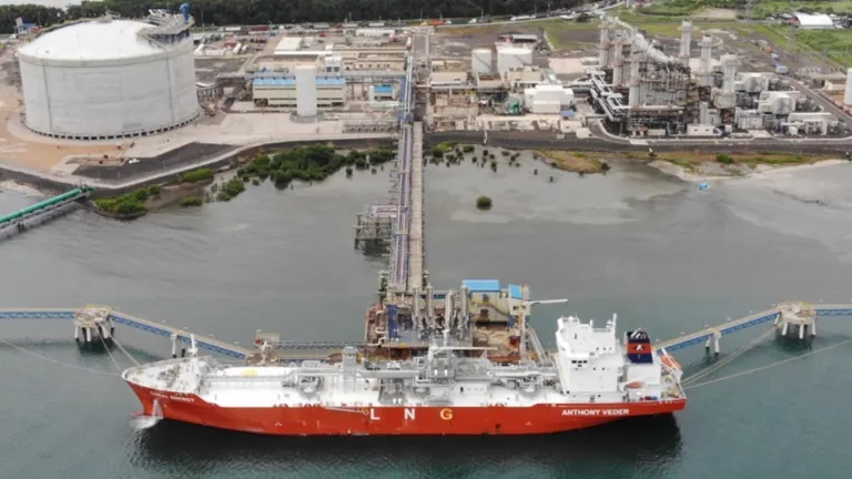 Aerial view of an LNG tanker docked at a large industrial terminal. The terminal features storage tanks, pipelines, and processing facilities surrounded by greenery. The tanker is moored at a jetty extending into the water.