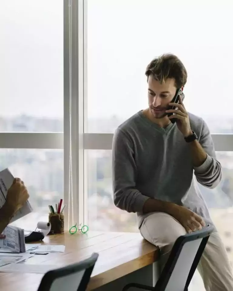 Two men in a windowed office talking; one is sitting on the desk on the phone.