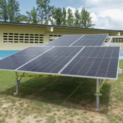 Paneles solares instalados en un campo con césped cerca de un edificio con una pared amarilla y azul, rodeados de cinta de precaución.