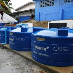 Tanques de almacenamiento de agua azules etiquetados 'Tanker+' están alineados al aire libre sobre una plataforma de concreto. Los tanques tienen una capacidad de 5000 litros y están situados cerca de un edificio azul.