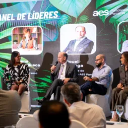 Cuatro panelistas sentados en el escenario durante una discusión de panel de liderazgo. Un hombre con micrófono habla mientras los demás escuchan. Una gran pantalla detrás muestra 'Panel de Líderes' con imágenes de los oradores. Miembros del público son visibles en primer plano.