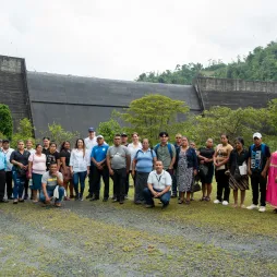 Group of people standing in front of a large dam surrounded by lush greenery and trees.