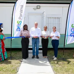Five people standing in front of a building holding a blue ribbon for a ribbon-cutting ceremony. Two women on the sides are wearing colorful traditional dresses. Two banners with 'AES Panamá' are displayed.
