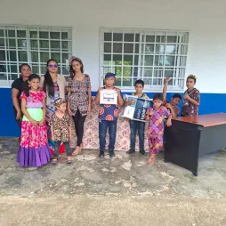 A group of children and adults stand in front of a building with white and blue walls. Some children are holding items, and they are all dressed in colorful clothing. A desk is visible on the right side.